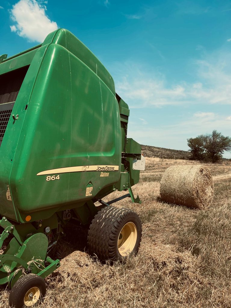 A green baler and hay bale under a bright blue sky, showcasing farming equipment and harvest.